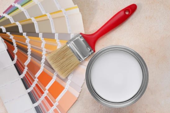 Scène d'intérieur avec pots de peinture ouverts disposés sur surface sombre, nuancier couleurs déployé en éventail, pinceau plat posé sur contenants